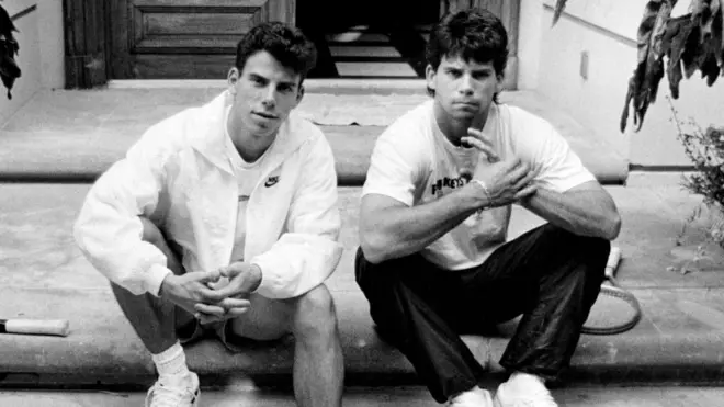 A foto em preto e branco mostra dois homens jovens, sentados no chão, um ao lado do outro, olhando para a câmera.