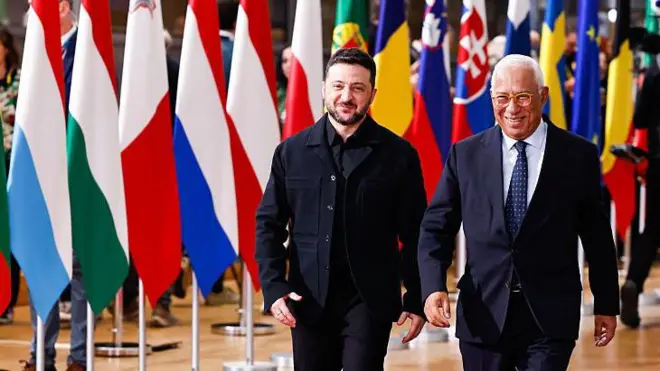 Владимир Зеленский и Антониу Кошта улыбаются, на заднем плане множество флагов стран-членов Еврособза