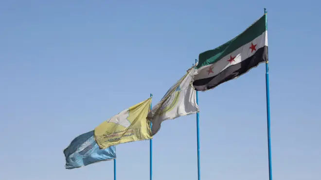 Sol baştan itibaren mavi, sarı, beyaz üç bayrak var. Sarı olan SDG bayrağı, beyaz olan Kuzey ve Doğu Suriye Demokratik Özerk Yönetimi bayrağı, mavi olan ise SDG'nin iç güvenlik sorumlu gücü Asayiş bayrağı. En sağda ise üç kırmızı yıldızlı ve yeşil, beyaz ve siyah şeritli Suriye bayrağı var.