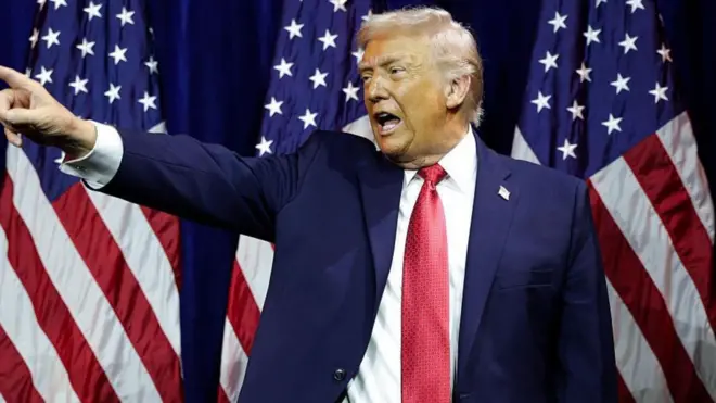 الرئيس ترامب يقف أمام علم الولايات المتحدة ويشير بيده اليمنى