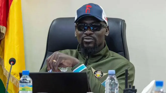 Photo en pied de Mamady Doumbouya vêtu d'un survêtement, d'une casquette de baseball et de lunettes noires.