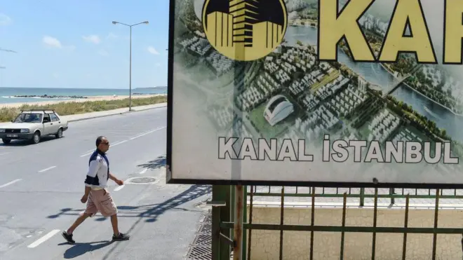 Kanal İstanbul çevresine yapılacak inşaatların çizimini gösteren bir pano, yanında bir erkek yürüyor, arkada deniz gözüküyor