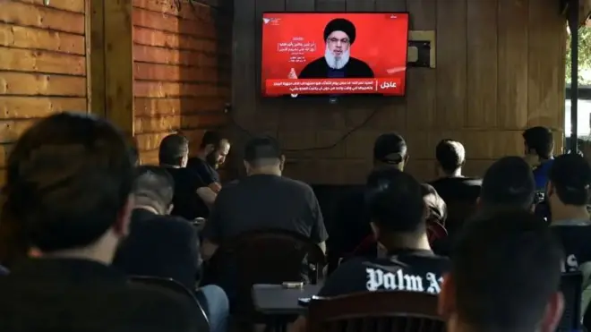 Foto of Hezbollah leader giving speech as pipo dey watch am from coffee shop