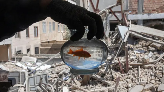 تهران، ایران – دهم فروردین: مردی یک ماهی قرمز را در تنگ آن در برابر آپارتمان آسیب دیده در حمله هوایی نشان می‌دهد