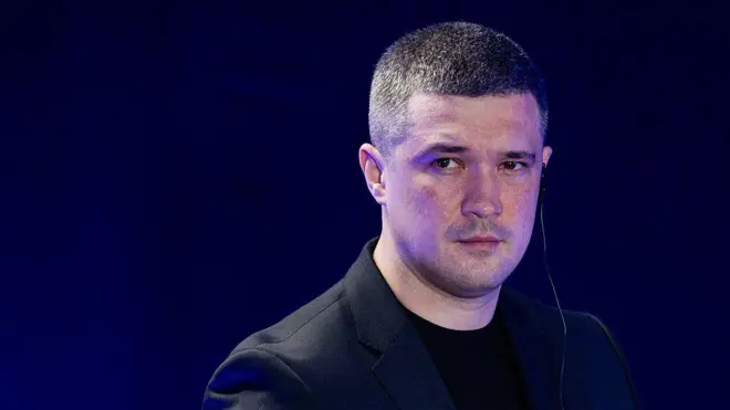 Портрет людини в темному піджаку та чорній футболці на тлі синього екрана. Людина стоїть перед камерою, на вусі видно навушник із тонким дротом. Знімок зроблений крупним планом під час публічного заходу або виступу.