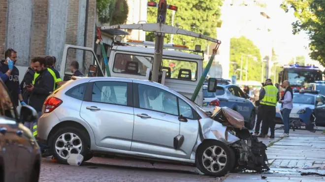 A silver car with the front mangled up next to a recovery vehicle, with police and police cars visible in the middle of the road