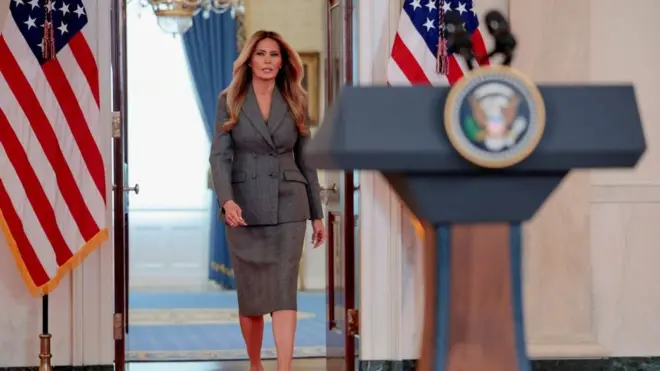 Melania Trump is wearing a business suit with a skirt as she walks up to a podium with the presidential seal