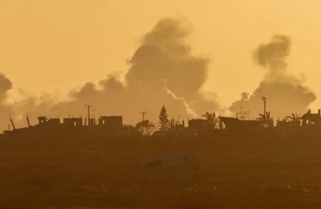 تصاعد الدخان من غزة بعد انفجار بالقرب من الحدود بين إسرائيل وغزة، كما شوهد من إسرائيل، الصورة تميل إلى اللون الأصفر. هناك القليل من الأشجار المتفرقة وبعض المباني صغيرة الحجم.