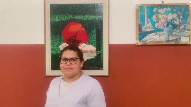 Edineusa com sorriso contido, em pé, em frente a parede com 2 quadros