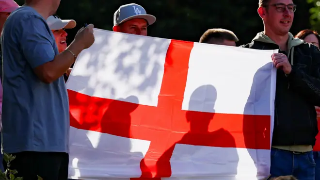 Трое мужчин держат английский флаг на протесте у гостиницы с мигрантами в Алтринчеме под Манчестером 8 августа 2025 года