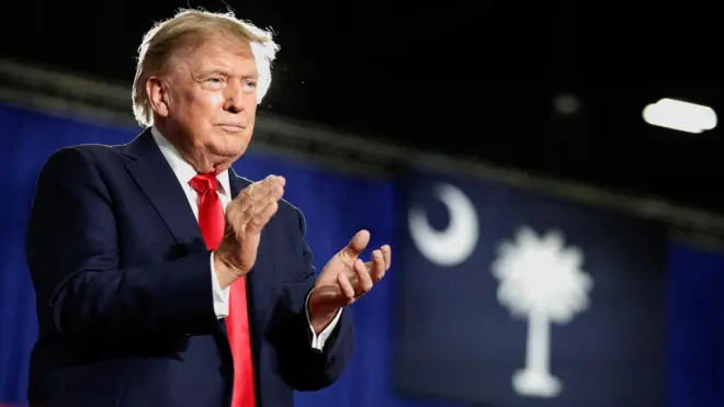 Perezida w'Amerika Donald Trump ahagaze ari ahavugirwa ijambo hegutse, yambaye ikositimu y'ubururu butsitse na karuvati itukura, arimo gukomera amashyi abantu batagaragara mu ifoto. 
