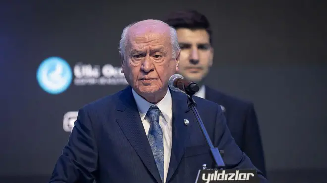 Bahçeli kürsüde lacıvert takım elbisesiyle ayakta konuşma yapıyor. Beyaz seyrek saçları ile sert bir duruşu var. Arkada bir erkek daha duruyor.