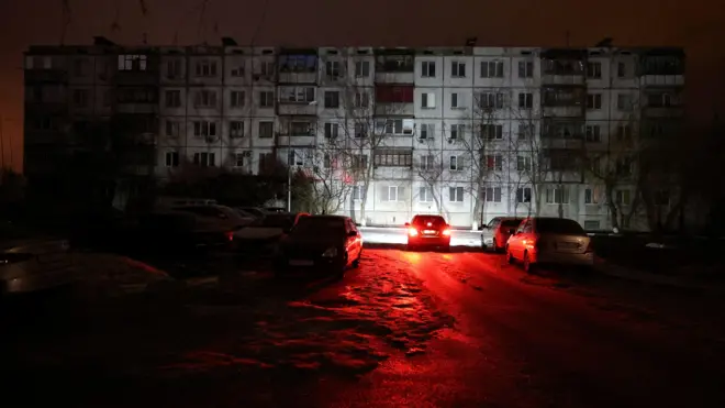 На заднем плане - обесточенный панельный пятиэтаный многоквартиный дом. На переднем плане дорога, освещенная красными фарами автомобиля перед домом.