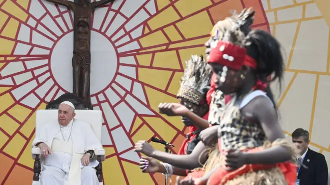 Le pape François (G) en habits blancs regarde la cérémonie d'accueil au stade des Martyrs à Kinshasa, en République démocratique du Congo. Il est assis devant un mur avec une croix en bois qui est montée sur un mur coloré avec des motifs blancs, jaunes et orange avec des lignes de mosaïque rouges. Des enfants vêtus de vêtements traditionnels dansent dans le cadre droit de la photo.