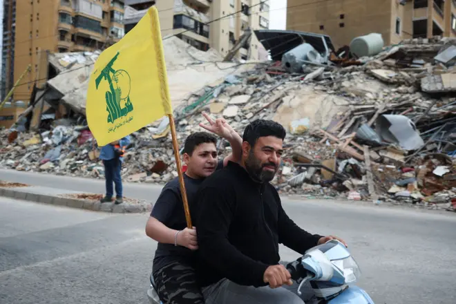 شخصان يستقلّان دراجة نارية صغيرة، يرفع أحدهما علم حزب الله ويشير بإصبعين في الهواء، في شارع في بيروت أمام كومة كبيرة من الركام حيث كان مبنى قائماً، في صورة التُقطت يوم الجمعة.

