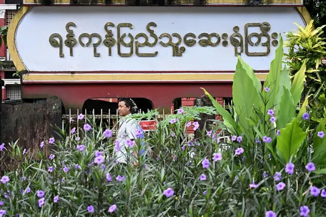 ပြီးခဲ့တဲ့သြဂုတ်လ ၁၇ ရက်တုန်းက မြင်ရတဲ့ ရန်ကုန်မြို့ ဆေးရုံကြီး မြင်ကွင်း၊ ရန်ကုန်ဆေးရုံကြီးလို့ မြန်မာလို ရေးထားတဲ့ဆိုင်းဘုတ်ကြီးနဲ့ အရှေ့မှာ အင်္ကျီအဖြူ၀တ်တဲ့အမျိုးသား တစ်ဦး ဖြတ်လျှောက်သွား၊ ပုံထဲမှာ ခရမ်းရောင်ပန်းပွင့်နေတဲ့အပင်စိမ်းစိမ်းအချို့ကိုလည်း မြင်ရ