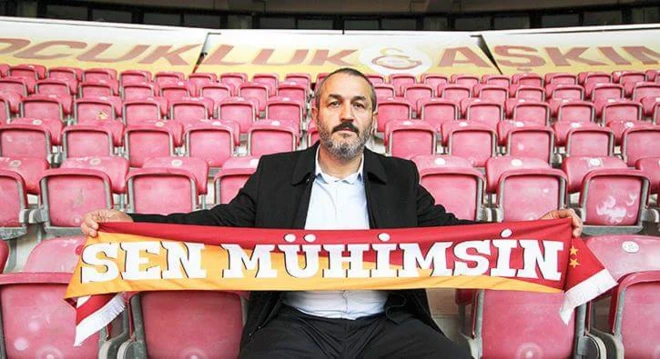 Sebahattin Şirin sarı kırmızılı arka plan üzerine "sen mühimsin" yazılı bir Galatasaray kaşkolunu tutuyor ve tribünde kırmızı koltuklar üzerinde oturuyor