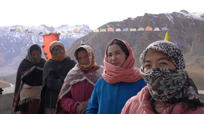 Five women wearing winter jackets look into the camera. They are standing in front of snow-covered peaks. 