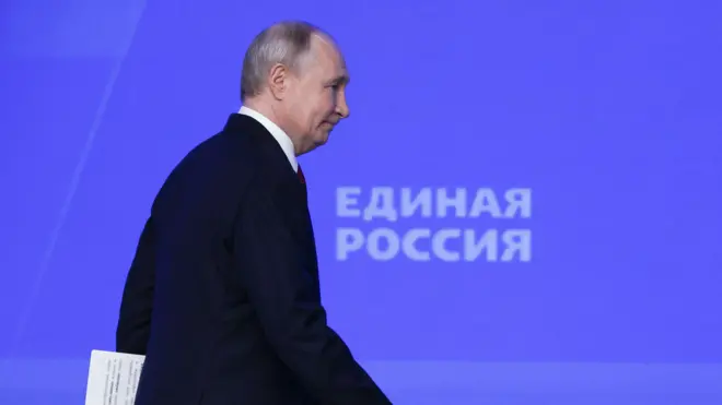 Путин в костюме с бумагами в руках идет по сцене на фоне баннера с надписью «Единая Россия». 
