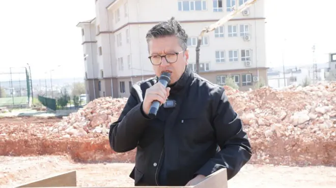 Şeref Albayrak bir inşaatın önüne kurulmuş kürsüde, elinde mikrofonla konuşurken. Üzerinde siyah bir mont var