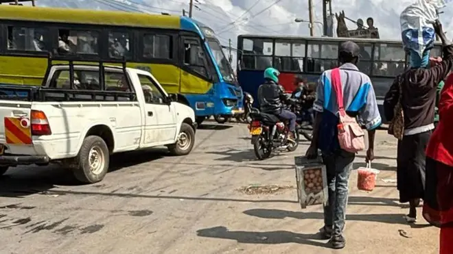 Umugabo akiri muto afashe mu kuboko kw'i bubamfu agasandugu k'amagi n'akadobo ka gachumbari mu kuboko kw'ukuryom, ahetse agasakoshi yacishije umugozi wako ku rutugu i bubamfu kenenera kw'ifyinga ry'i buryo, imbere yiwe hari abamotari n'umugore yiremetse umufuko, i bubamfu bwiwe haboneka kamyoneti n'ibisi nini 