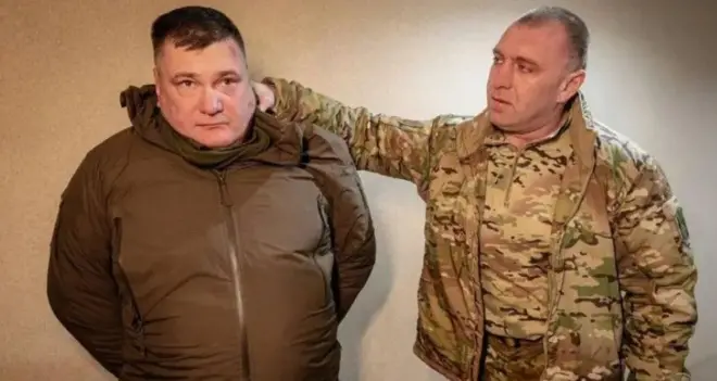 Малюк у кутрці кольору хакі тримає руку на шиї чоловіка у коричневій куртці. У чоловіка руки за спиною 