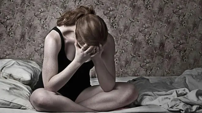 Mujer sentada en una cama, con la cabeza entre sus manos. Tonos opacos