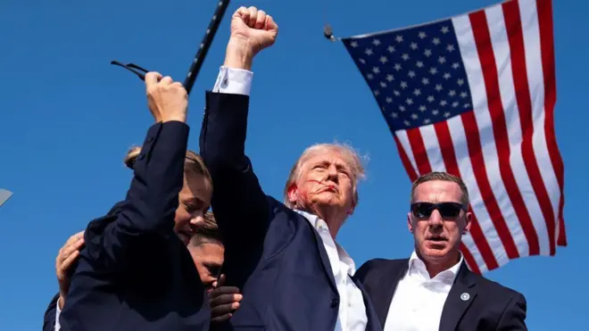 Donald Trumppagbet fora do arpunho cerrado e rosto ensanguentado cercado por agentespagbet fora do arsegurança com bandeira dos EUA ao fundo