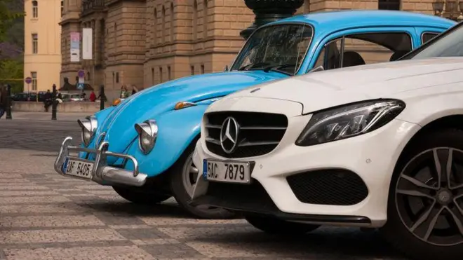 Um pequeno Fusca azul da Volkswagen ao lado de um grande Mercedes branco, estacionados em Praga, na República Tcheca
