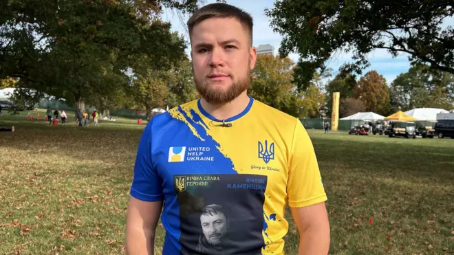 Dmytro Kamenschyk com camiseta azul e amarela, cores da bandeira da Ucrânia, e a foto do pai deleflamengo e america mg palpitepreto e branco