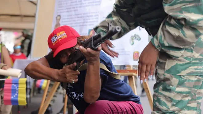 Una mujer vestida de civil con una gorra a favor del gobierno de Maduro apunta un arma teniendo una de sus rodillas apoyadas en el piso. Otra mujer, cuyo rostro no vemos pero que lleva un uniforme militar y uñas pintadas le ayuda a apuntar. En el fondo, se ven banderas venezolanas.