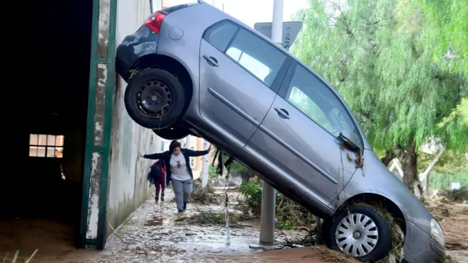 A car with its r=ear wheels high up against a wall and its front pointing into mud while a woman can be seen beneath its rear wheels as she walks towards the vehicle