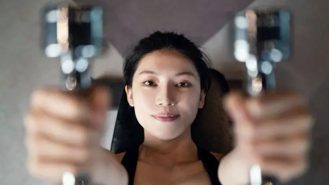 A woman holds weights handles on either side of her face, with the handles out of focus clpose to camera and her face in perfect focus