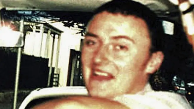 A man with short, dark hair is sitting with his arm across the back of a car seat