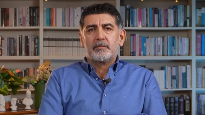 Gültekin bir çalışma odasında kamera karşısında mavi gömleğiyle oturuyor arkasında kitaplık ve kitaplar var. 