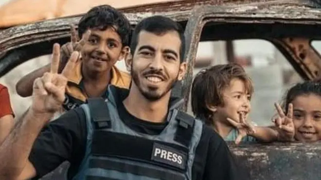 يبتسم الجعفراوي رافعاً علامة النصر ويرتدي سترة الصحفيين وخلفه عدد من الأطفال يأخذون صورة معه.