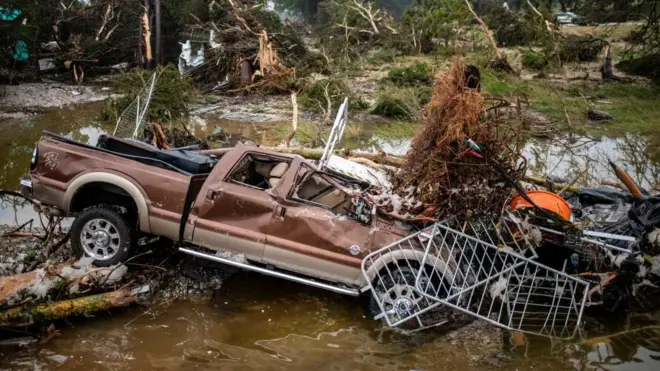 川の氾濫で押し流され車体がゆがんだ茶色いピックアップトラックが、フェンスや流木などにからまって泥の中で斜めになっている