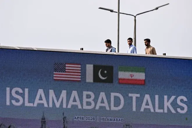 Большая надпись Islamabad talks, над ней флаги США, Пакистана и Ирана, сверху над билбордом проходят трое мужчин