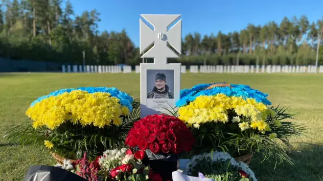Могила на кладовищі, білий хрест із фото, біля могили живі квіти червоні та жовто-блакитні