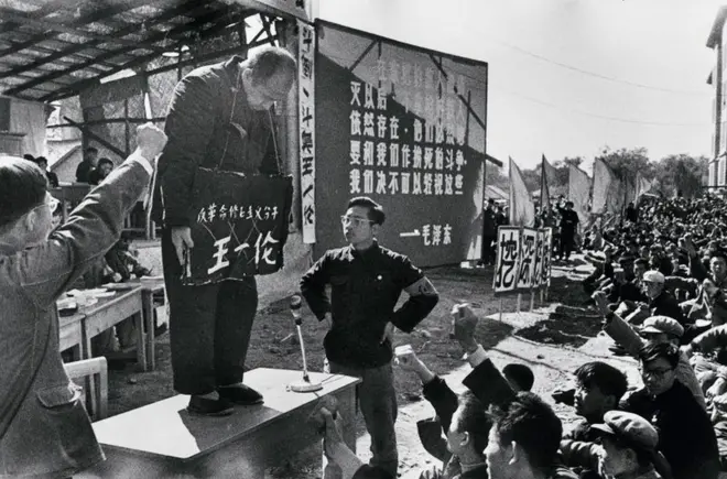 文革批鬥現場，一名長者站在桌上，頸掛一個寫有其姓名的牌子，周圍有群眾握拳。