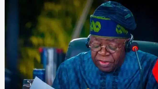 Aarẹ Naijiria, Bola Tinubu ninu aworan yii n wo isalẹ, iwe lo jọ pe o n ka lọwọ. Aarẹ wọ aṣọ alawọ aro, o de fila alawọ aaro o si fi igo soju. Ẹrọ gbohun-gbohun n tọka sibi eti rẹ, bẹẹ lo lo ẹrọ ti eeyan fi maa n da nikan gbọrọ si eti rẹ mejeeji.