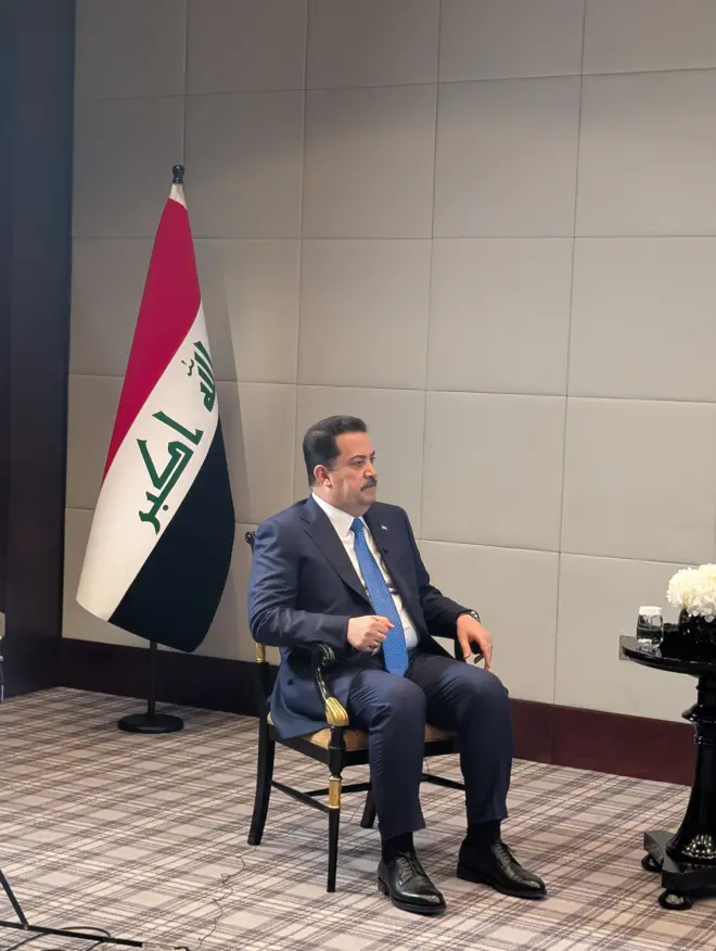 رئيس الحكومة العراقية محمد شياع السوداني في مقابلة مع بي بي سي، يرتدي بدلة رسمية كحلية وقميص أبيض وربطة عنق باللون الأزرق وخلفه علم العراق.