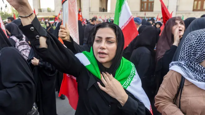 テヘランで、イランの国旗を肩に巻いた若いイラン人女性が、旗を振る人々の群衆の中で腕を高く掲げて口を開いている