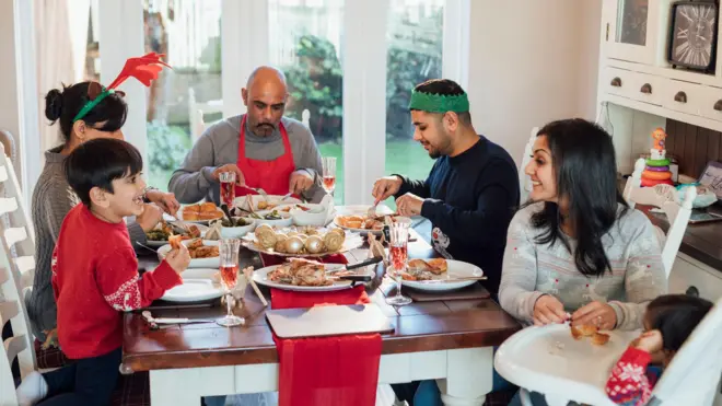 A multi-generation family sitting in the dining room eating Christmas Dinner together. One woman is helping her son eat while he is sitting in a high chair.