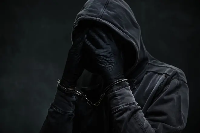 Criminel menotté portant un sweat à capuche et couvrant le visage avec les mains.