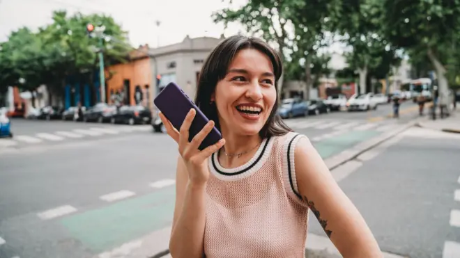 una mujer en medio de una calle envía una nota de voz. sonrié y tiene el celular en la mano. 