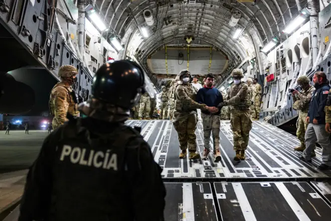 Двое военных с закрытыми масками лицами ведут мужчину по трапу, его встречает полицейский.