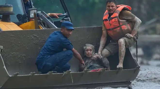 На фото - рятувальна операція після масштабної повені. Двоє чоловіків за допомогою ковша будівельної техніки евакуюють літню жінку через затоплену та вкриту густим шаром бруду місцевість.