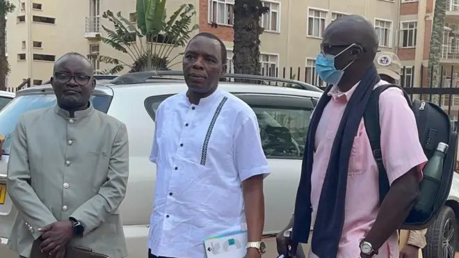 Jean Paul Micomyiza (iburyo), yambaye imyenda y'ibara rya roza iranga imfungwa, yambaye agapfukamunwa n'indorerwamo, fulari y'umukara mu ijosi, ahetse n'igikapu cy'umukara mu bitugu, yambaye n'isaha y'urushinge ku kuboko kw'ibumoso. Ahagararanye n'abagabo babiri bambaye imyenda isanzwe, bahagaze imbere y'imodoka y'umweru. Bari i Huye mu majyepfo y'u Rwanda. Ifoto yo ku itariki ya 13 Gicurasi (5) mu 2025. 