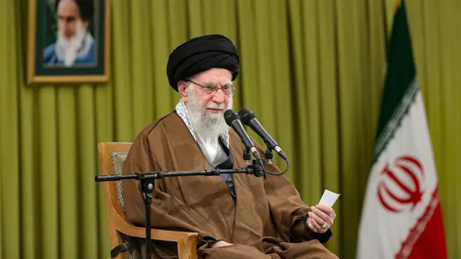 Hamaney bir koltukta oturuyor. Önünde mikrofon. Arkasında İran bayrağı ve Humeyni'nin fotoğrafı var.